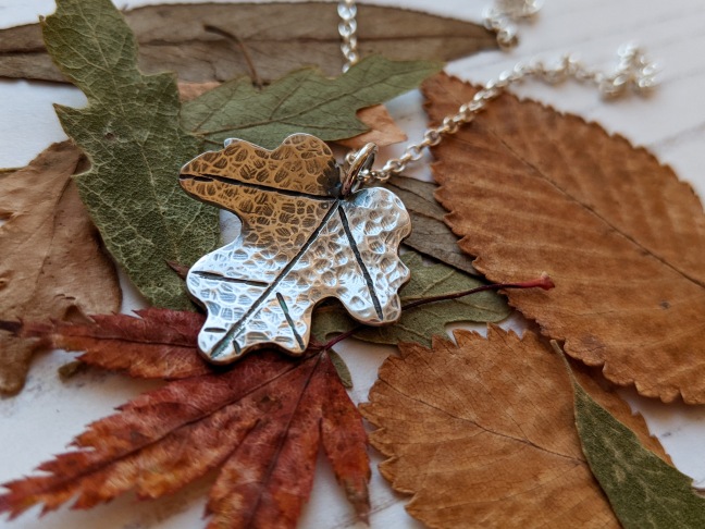 A maple leaf pendant formed from silver, with a silver chain, on a background of autumn coloured leaves
